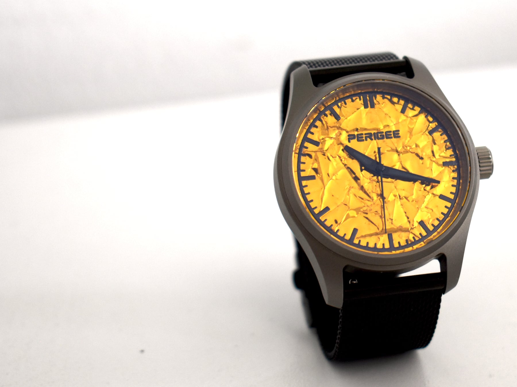 The PERIGEE Contact Light photographed on a white sweep: a 39 mm Grade 2 titanium case with a gold foil dial, dark baton markers and a black strap.